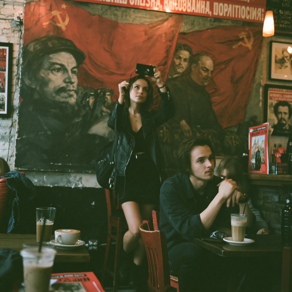 A young woman standing on her chair to photograph her latte from above while her companion waits